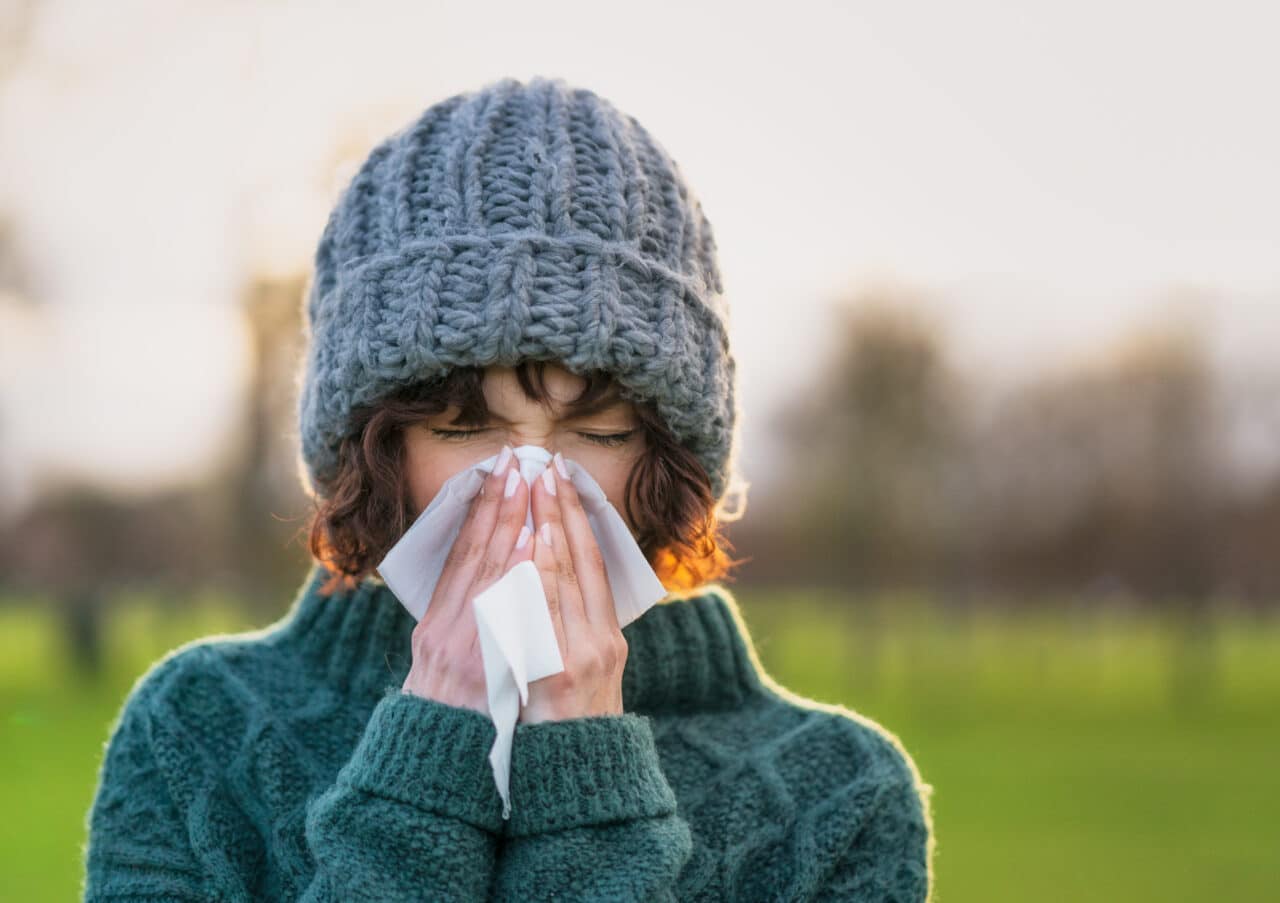 Woman wearing a warm hat and sweater blowing her nose. It is a chilly winter day.