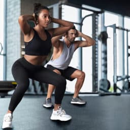 Two fit adults doing squats at a gymnasium. Staying physically active is good for hearing health on top of overall health.