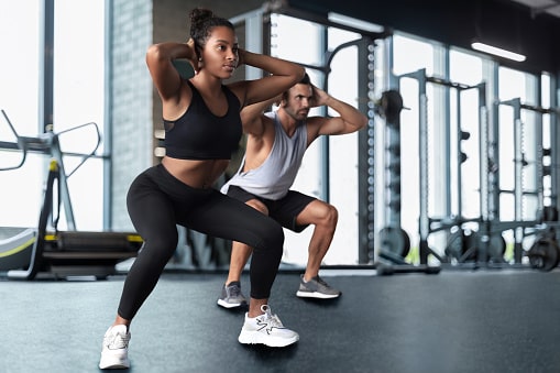 Two fit adults doing squats at a gymnasium. Staying physically active is good for hearing health on top of overall health.