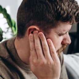 Man 30s touching ear, suffering from sudden throbbing ear ache, looking aside. Upset bearded male feeling unwell sitting on couch in living room at home