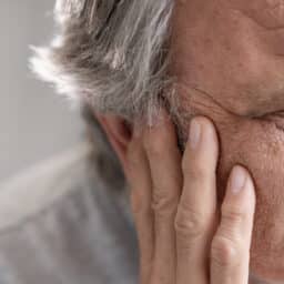 Man looking to left, head in full frame of image. Older with grey hair, holding right side of face with an expression of pain and distress